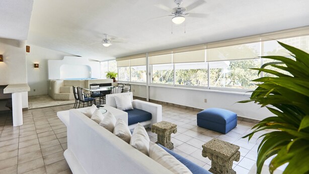A spacious living area with a sofa, blue ottoman, and a dining table with chairs. There are ceiling fans, large windows, and a potted plant. - Hotel Suites Ixtapa Plaza (Ixtapa)
