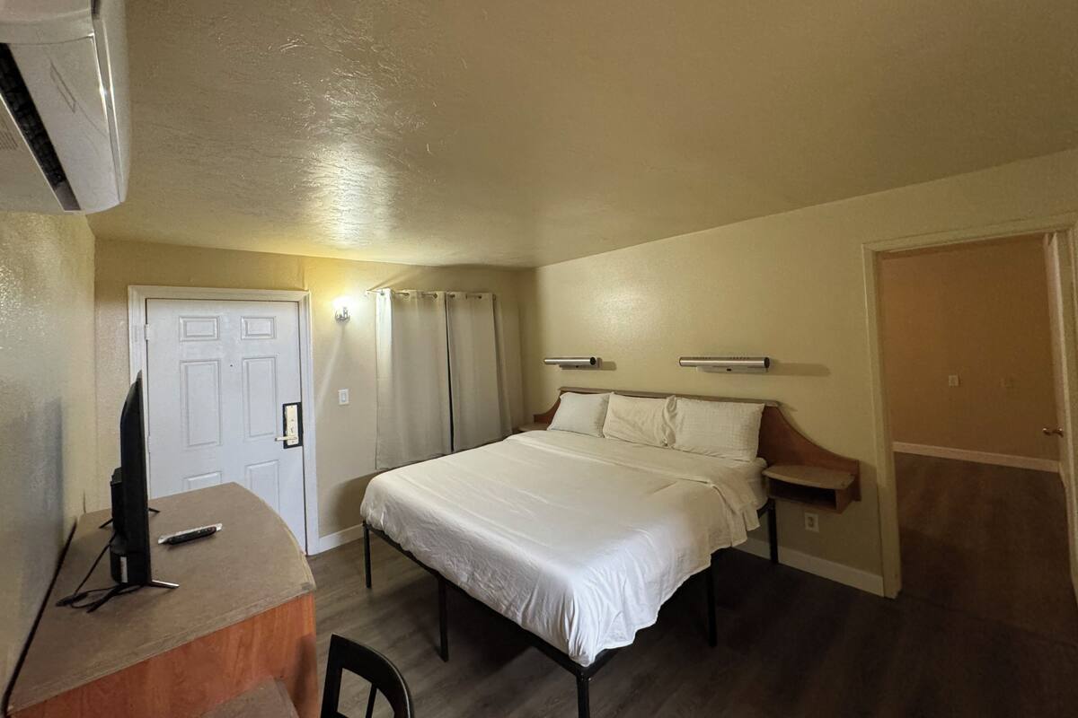 A bedroom with a single bed, a desk, and a chair. There is a door, a window with curtains, and a wall-mounted air conditioning unit.