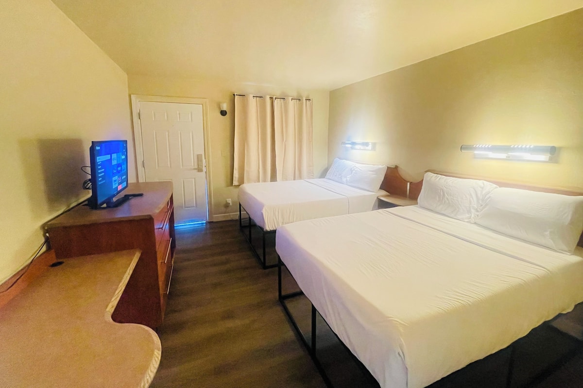 A hotel room with two beds, a desk with a computer, a door, and a light fixture on the wall.