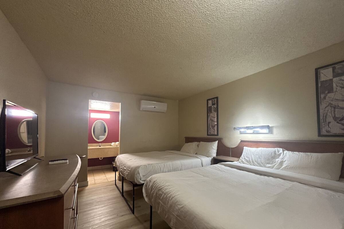 A hotel room with two beds, a desk, a mirror, a TV, and a wall-mounted air conditioner.