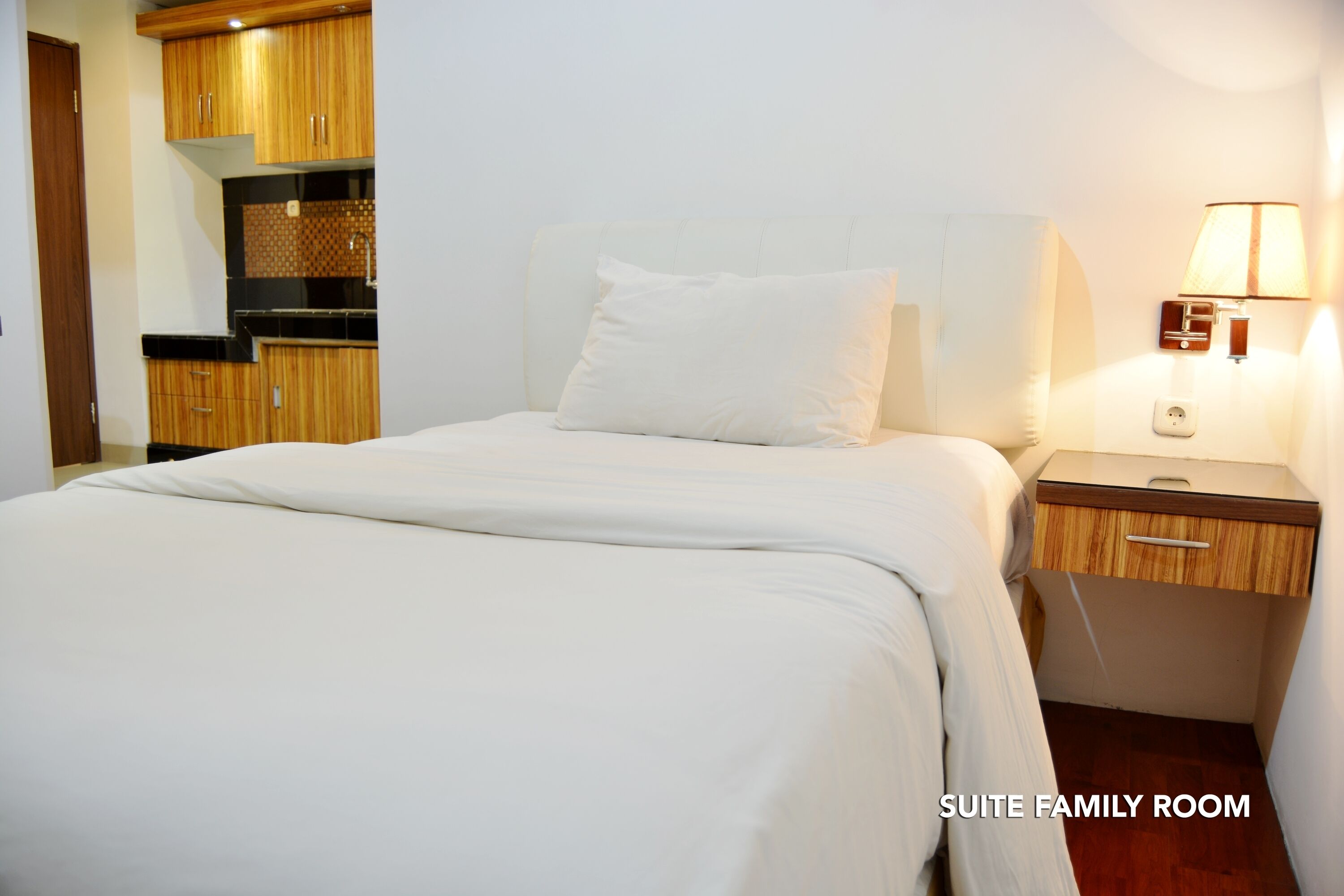 A suite family room with a bed, a nightstand, a lamp, and a kitchen area.