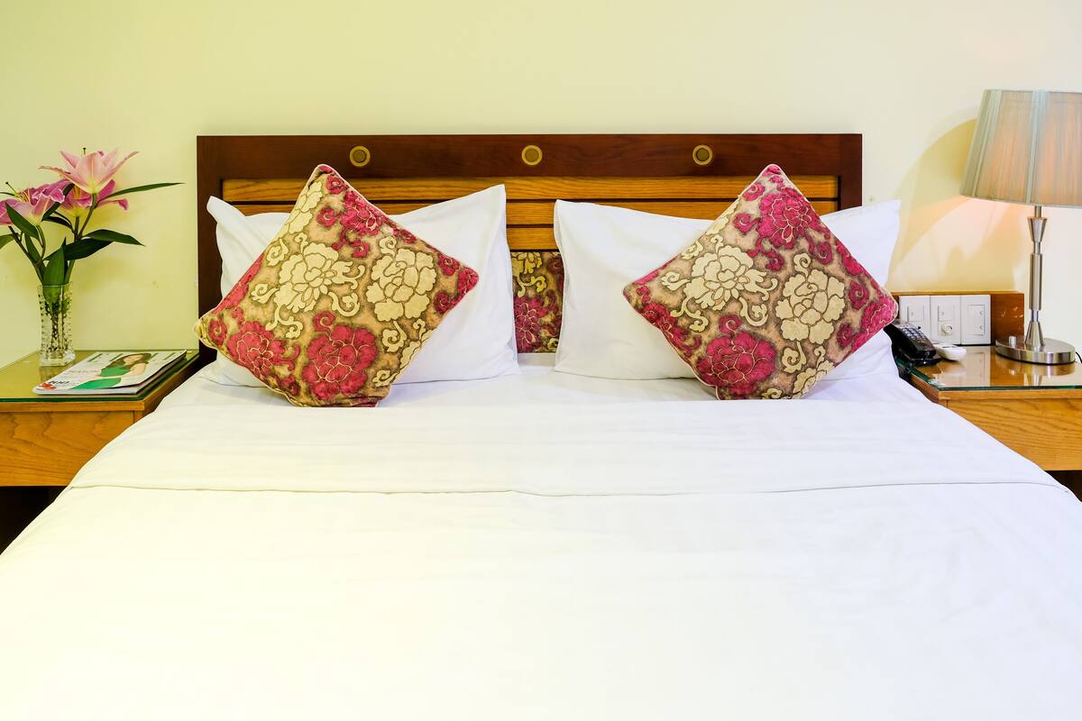 A neatly made bed with floral-patterned pillows, a bedside table with a lamp, and a vase with flowers.