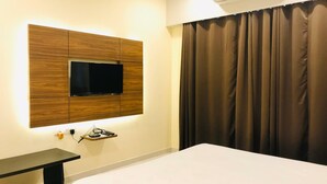 Et moderne hotelværelse med et fladskærms-tv monteret på en væg af træpaneler, et sengebord, en seng og et vindue med mørke gardiner.