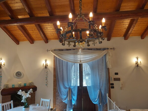 Ein Zimmer mit Holzbalkendecke, einer Chandelüster, einem Esstisch mit Stühlen, einem Kamin und einem fenster mit Vorhängen.