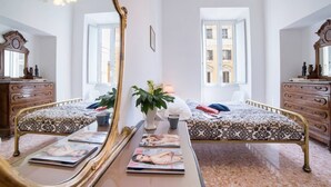 A bedroom with two beds, a large mirror, wooden furniture, and a view of buildings through the windows.