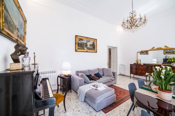 Una sala de estar con un piano, un sofá gris, una mesa de comedor con sillas, una araña y un cuadro en la pared.