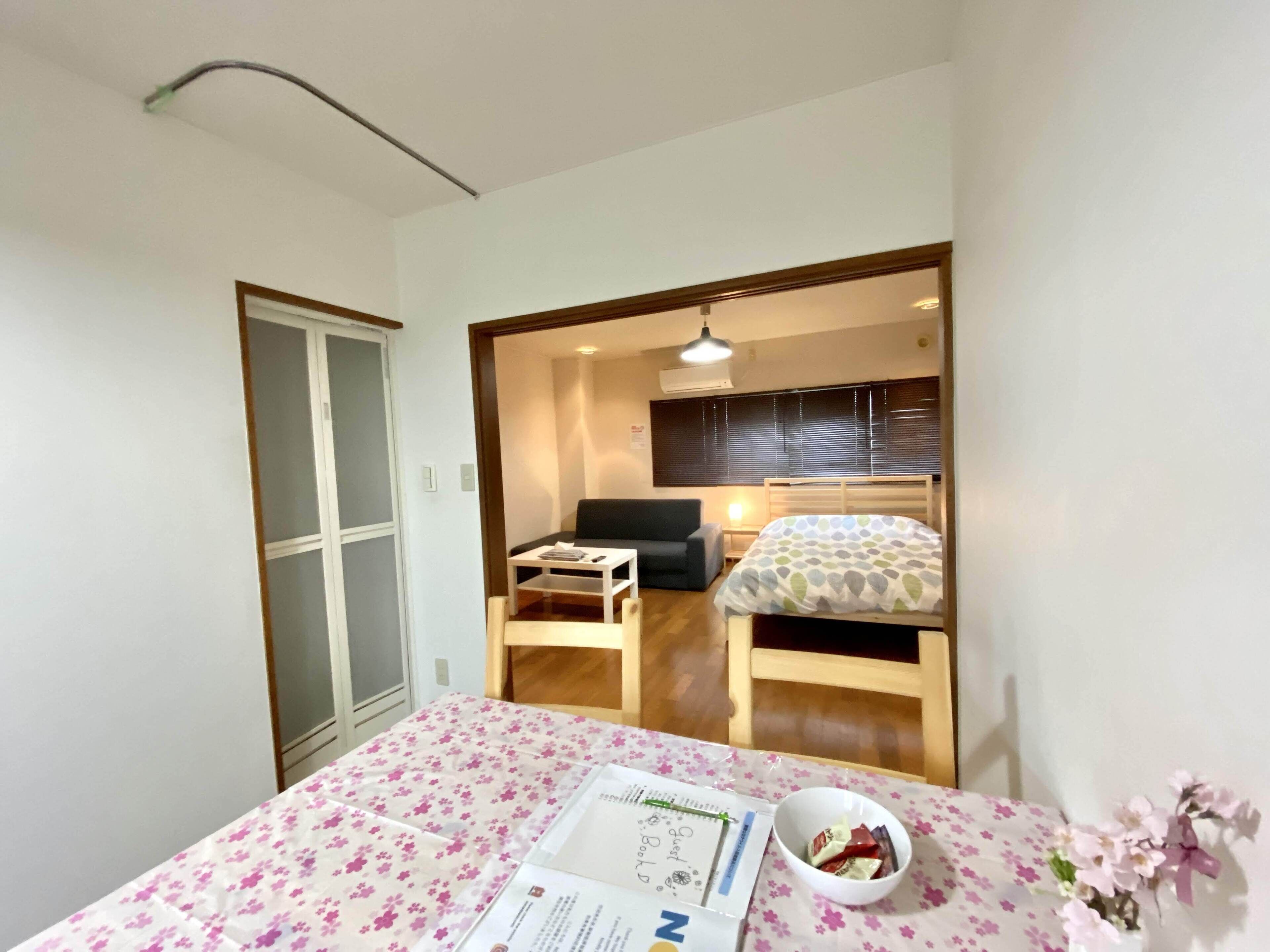 Habitación pequeña y bien iluminada con una cama, un escritorio y una silla. Cuenta con una ventana con persianas, una puerta y una mesita con un frutero.