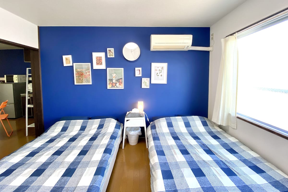 Una habitación con dos camas, kitchenette, ventilador de techo y ventana con cortinas.