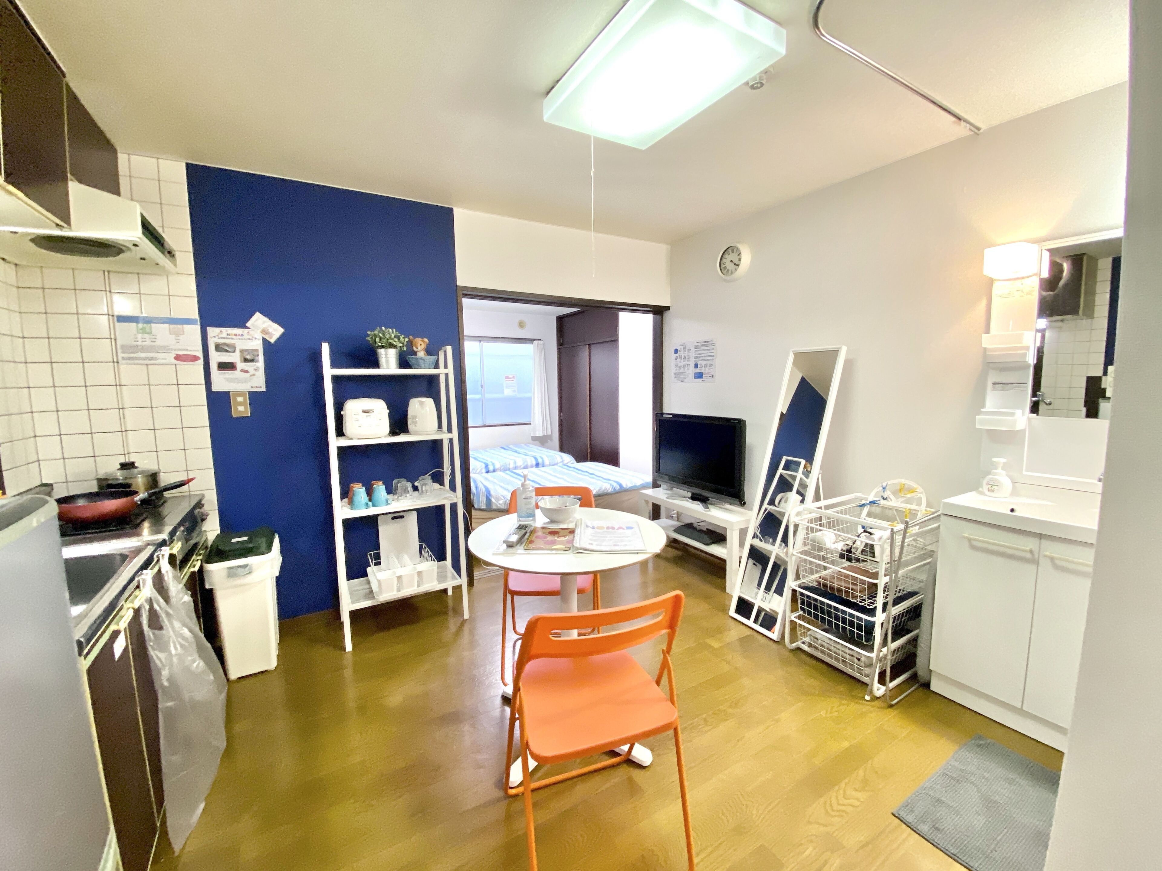 A compact hotel room with a kitchenette, a dining area with a table and chairs, a TV, a bed with a window, and a bathroom with a mirror and sink.