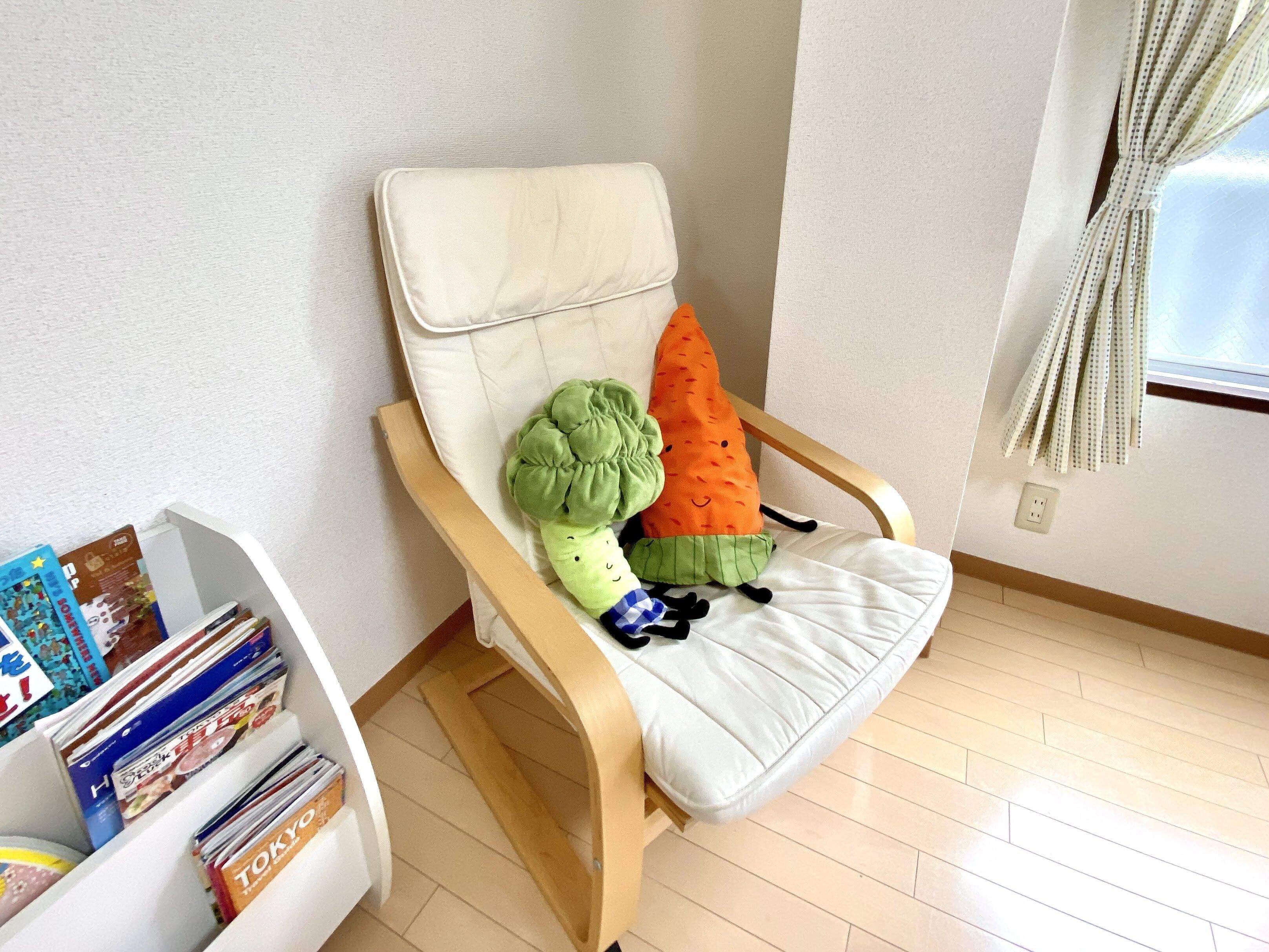A chair with a white cushion and two vegetable-shaped pillows, a bookshelf with magazines, and a window with curtains.