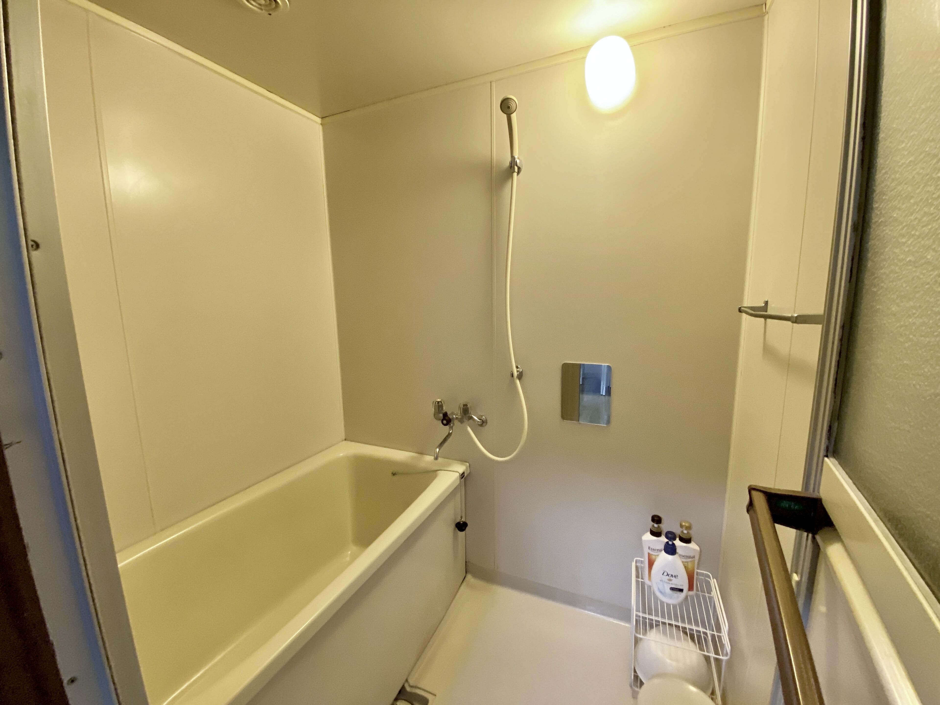 Une petite salle de bain blanche, équipée d’une baignoire, d’une douche et de toilettes. On y trouve une étagère avec des articles de toilette.