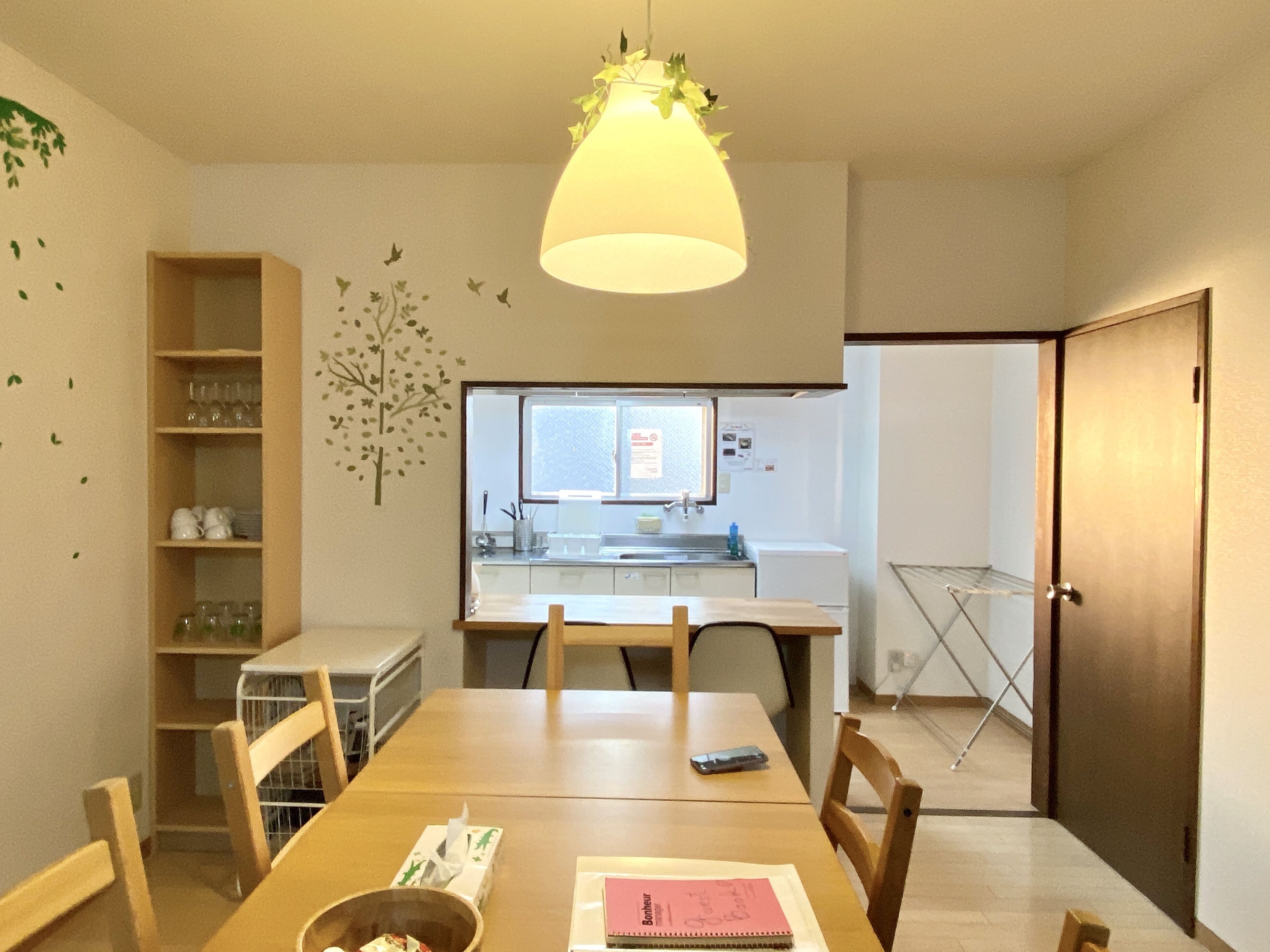 A dining area with a wooden table, chairs, a hanging light fixture, a window with a view of a kitchen, and a door.