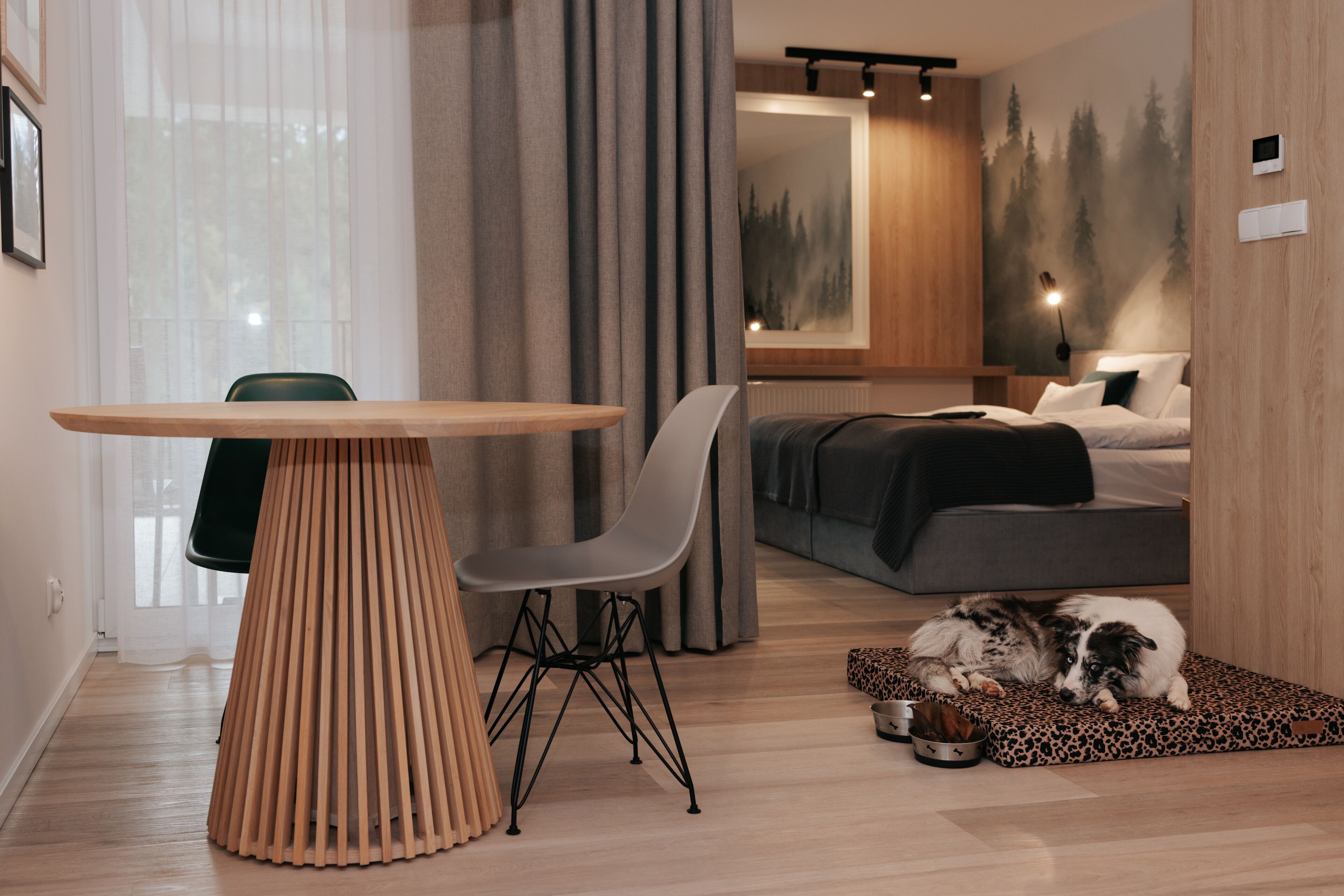 A modern living room with a wooden dining table, a sofa, a dog bed, and a large window with curtains.