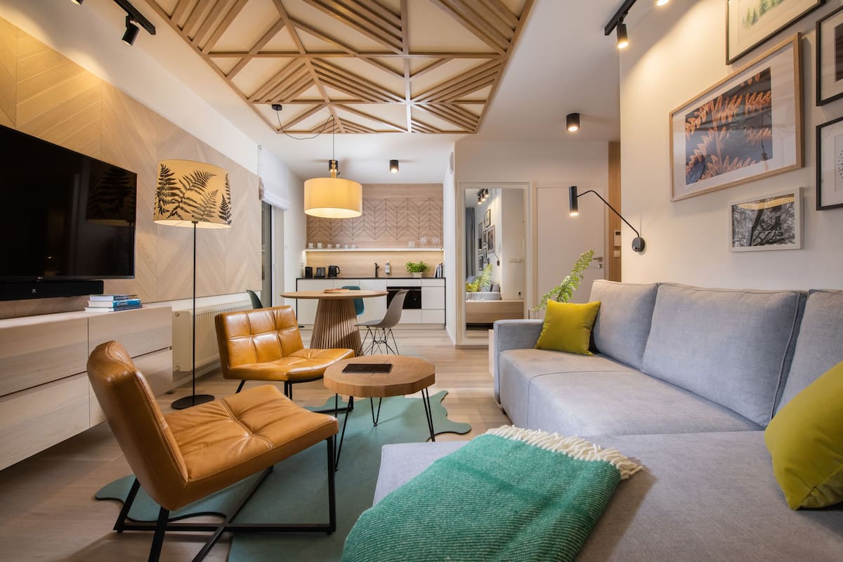 Una sala de estar moderna con un sofá, sillones y un televisor. Se observa una cocina al fondo con una mesa de comedor y sillas.