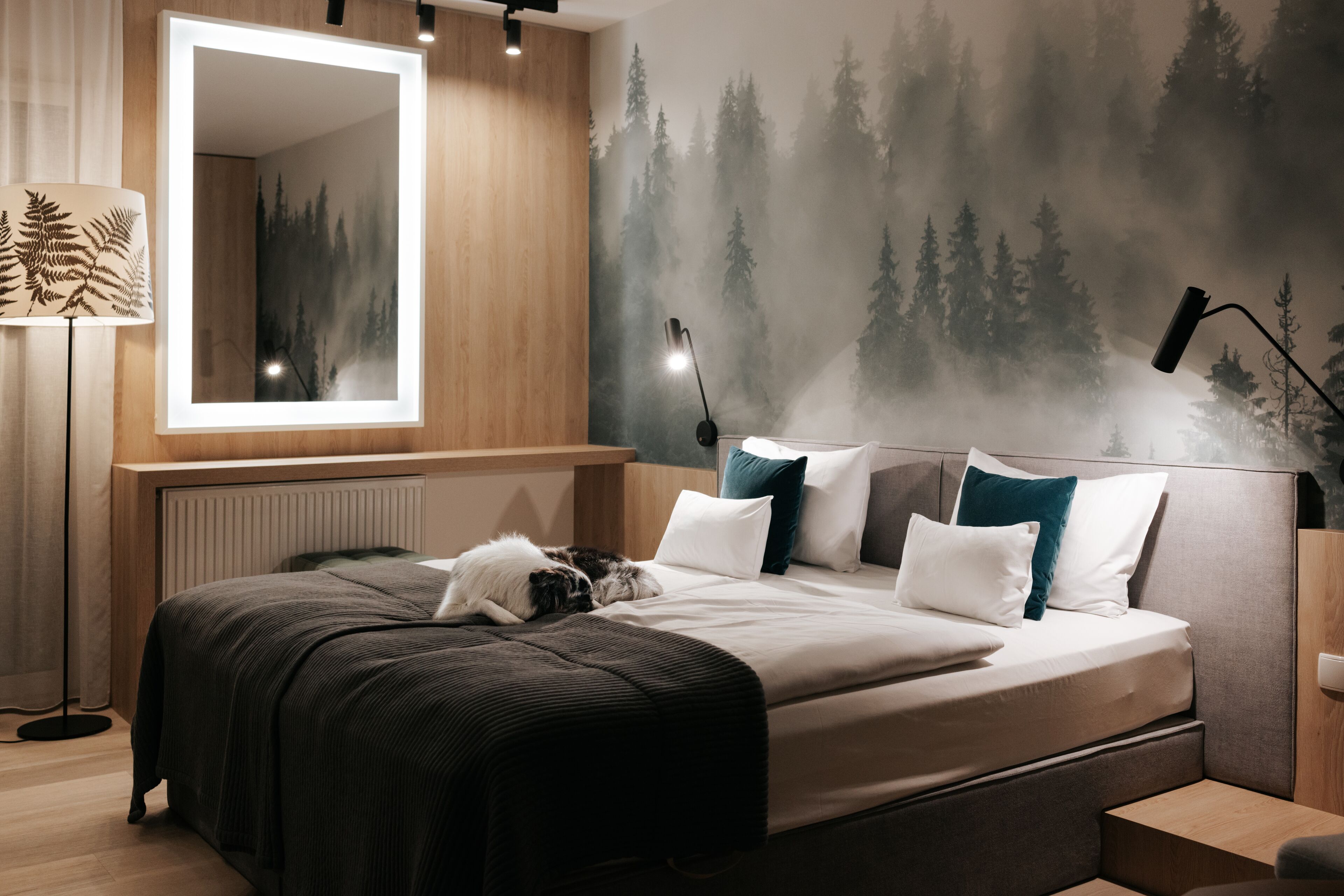 A modern bedroom with a large bed, a mirror, and a forest-themed wall mural.