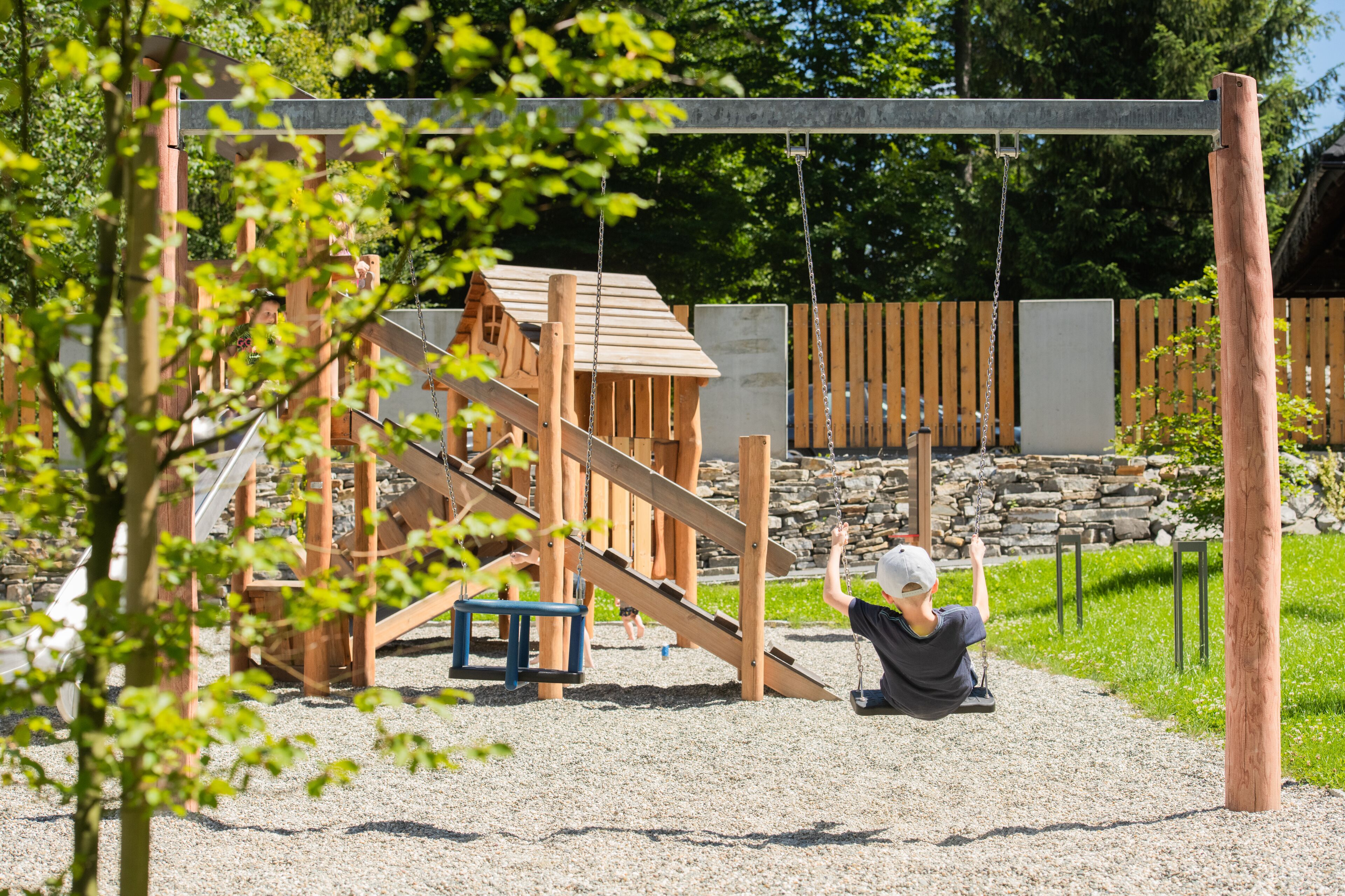 A playground with wooden structures, including a slide and swings, surrounded by gravel and greenery.