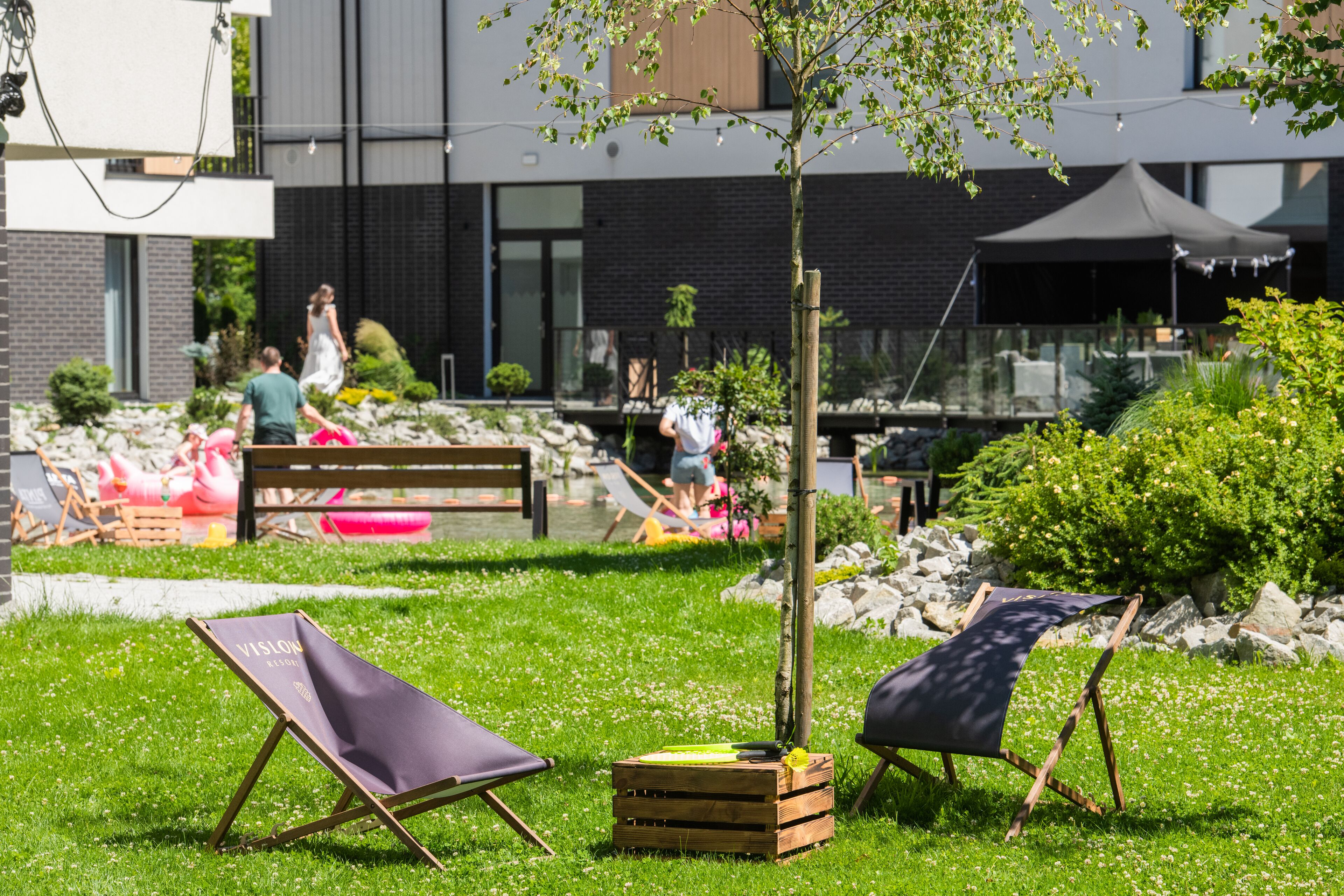 An outdoor seating area with wooden chairs and a small table, surrounded by greenery and a building in the background.