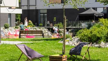 An outdoor seating area with wooden chairs and a small table, surrounded by greenery and a building in the background.