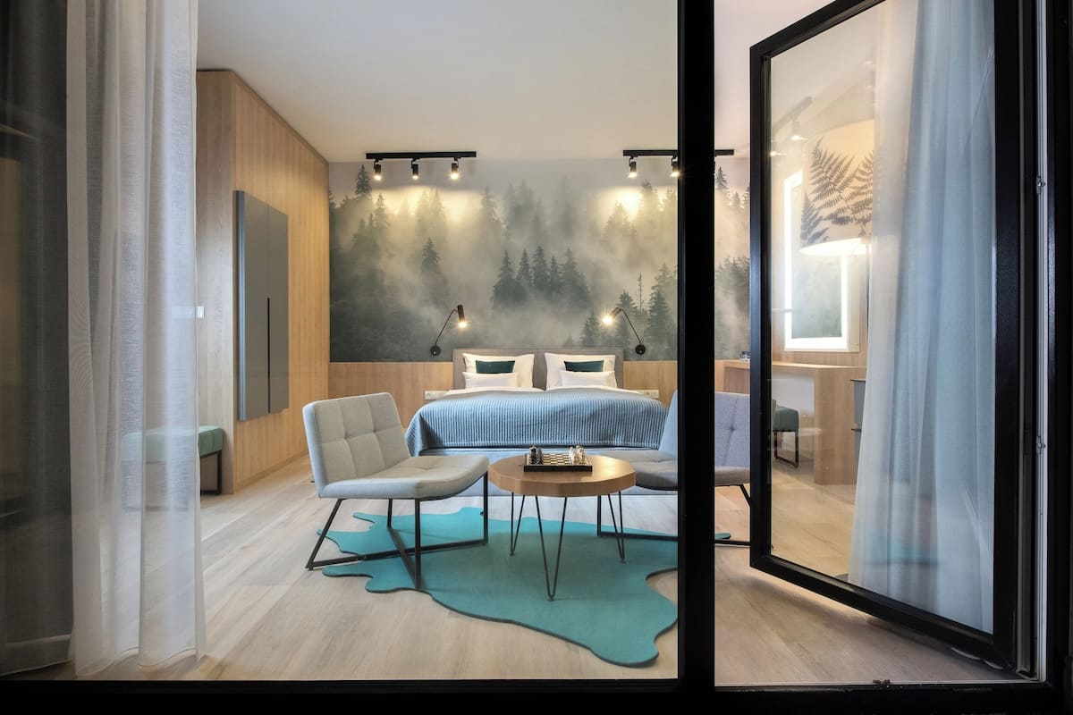 Un dormitorio moderno con cama, zona de estar con mesa y un amplio ventanal con vista a un bosque.