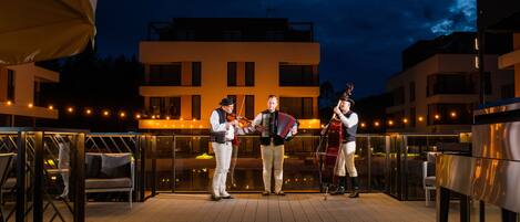 Une terrasse sur le toit, avec un bâtiment moderne en arrière-plan, une guirlande lumineuse et un groupe de personnes jouant d’instruments de musique.
