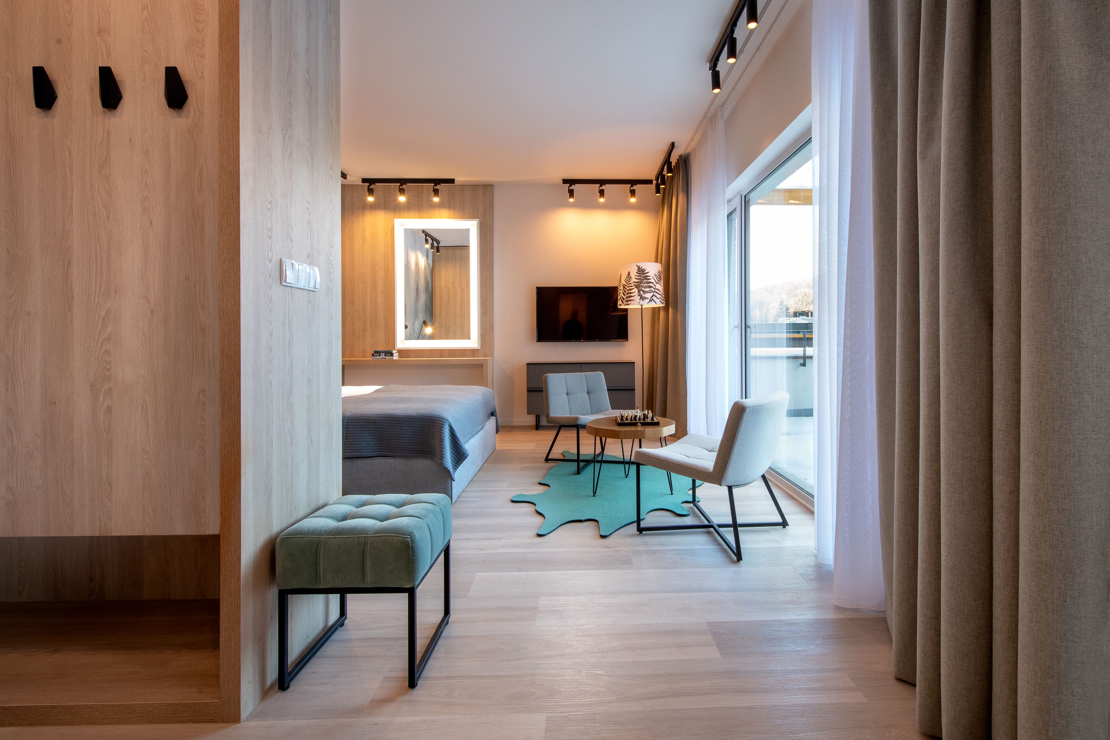 Ein modernes Hotelzimmer mit Holzboden, einem Bett, einer Sitzecke mit Tisch und Stühlen, einem großen Fenster mit Vorhängen und einem wandmontierten Flachbildfernseher.