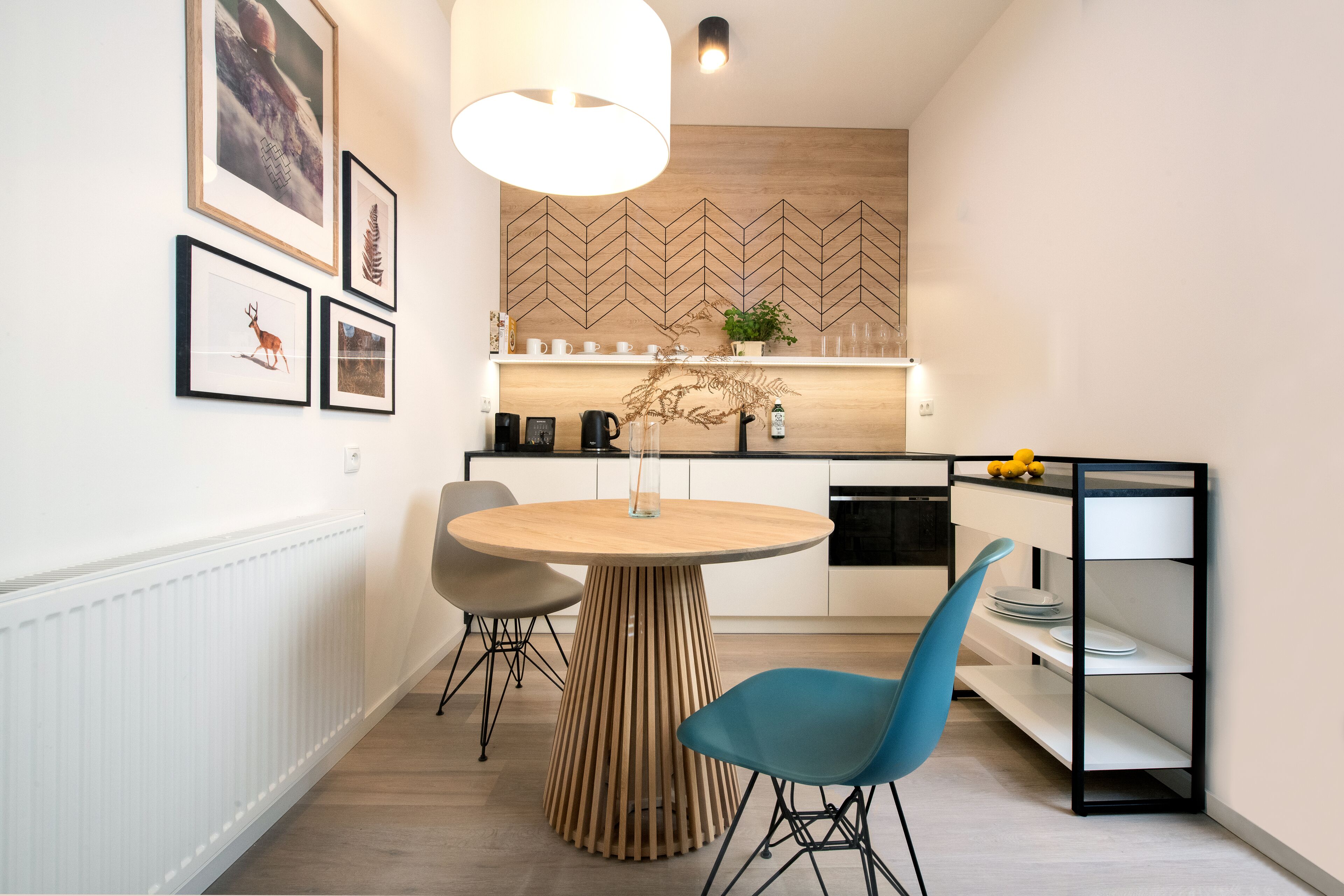 A modern kitchen with a round wooden table, two chairs, a hanging light fixture, and framed pictures on the wall.