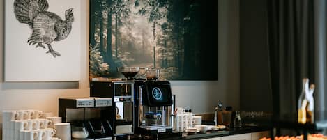 Una estación de café con una cafetera, tazas y un cuadro grande con temática forestal en la pared.