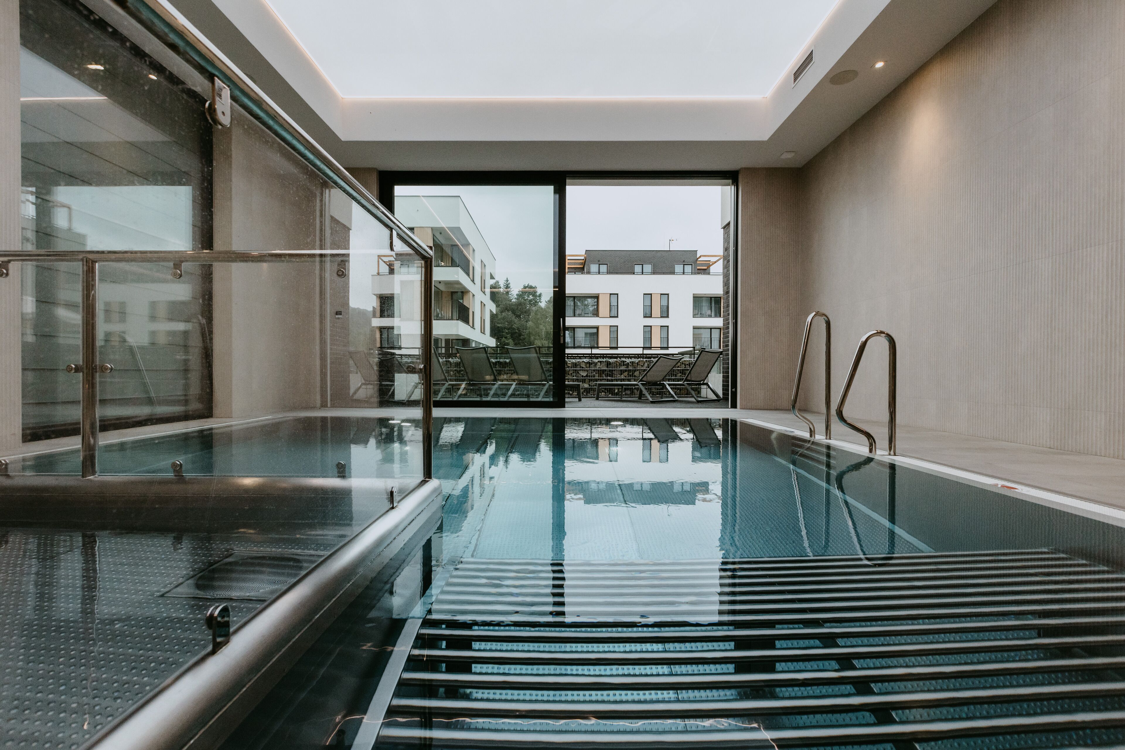A modern indoor swimming pool with glass walls, a clear view of the outside buildings, and a ceiling light fixture.