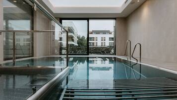 A modern indoor swimming pool with glass walls, a clear view of the outside buildings, and a ceiling light fixture.