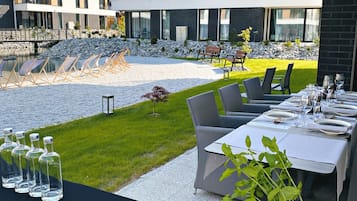 Un comedor al aire libre moderno con mesas dispuestas para una comida, un pequeño jardín y vistas a un edificio de varios pisos.