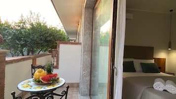 Un balcon avec une table dressée et ornée de fruits, un lit parfaitement fait et une porte-fenêtre coulissante donnant sur une pièce.