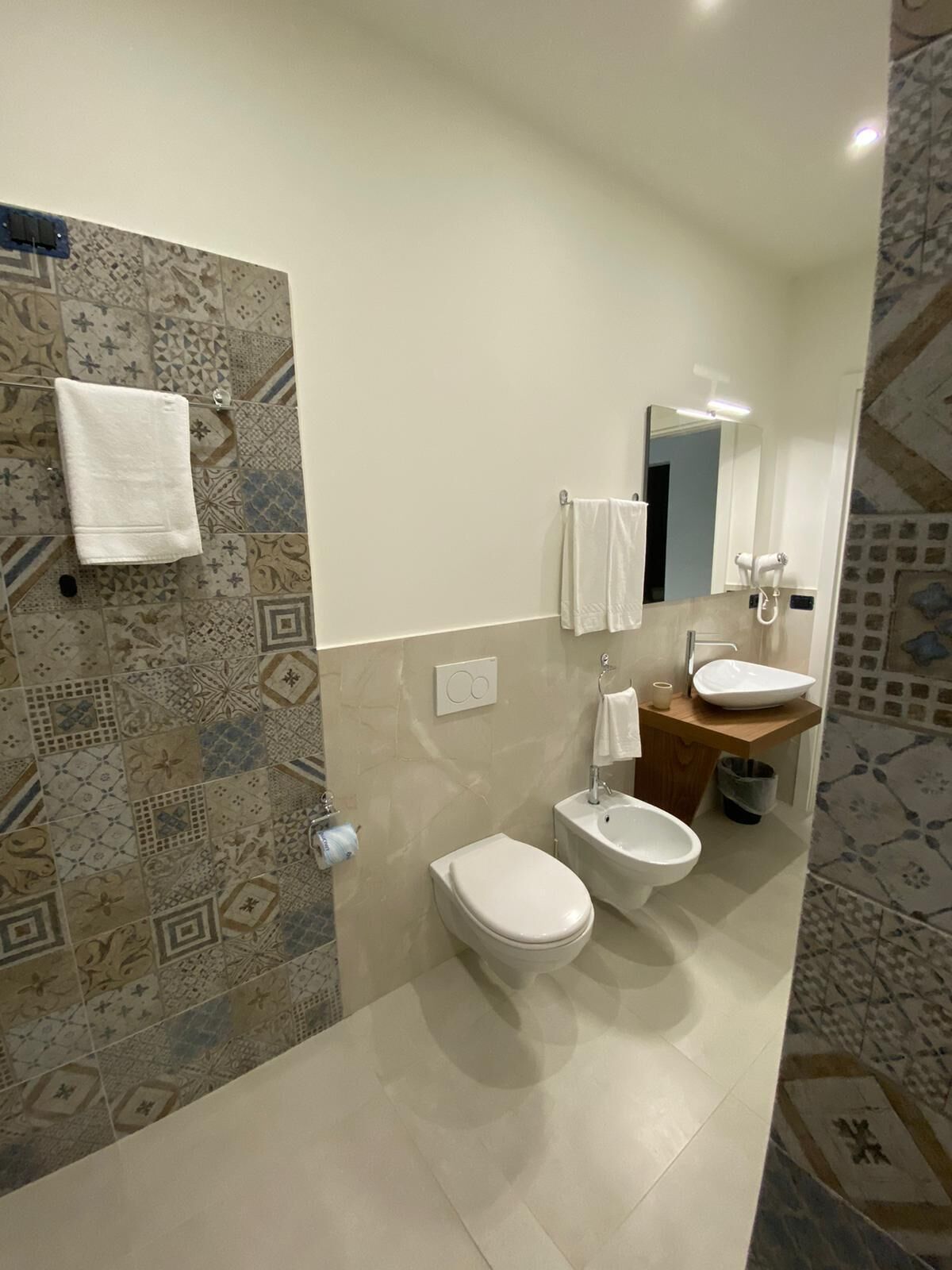 A modern bathroom with patterned tiles, a white toilet, a bidet, a sink, and a mirror.