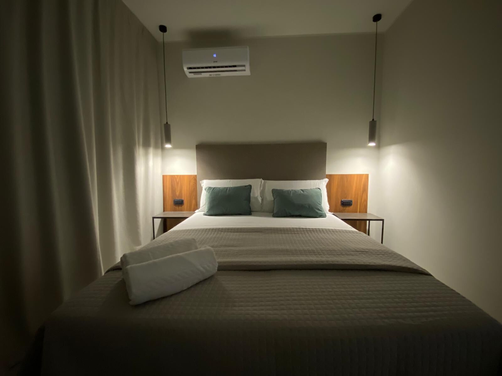Una cama bien hecha con un almohadón blanco y dos almohadones color turquesa, un cabecero de madera y dos mesitas de noche con lámparas.
