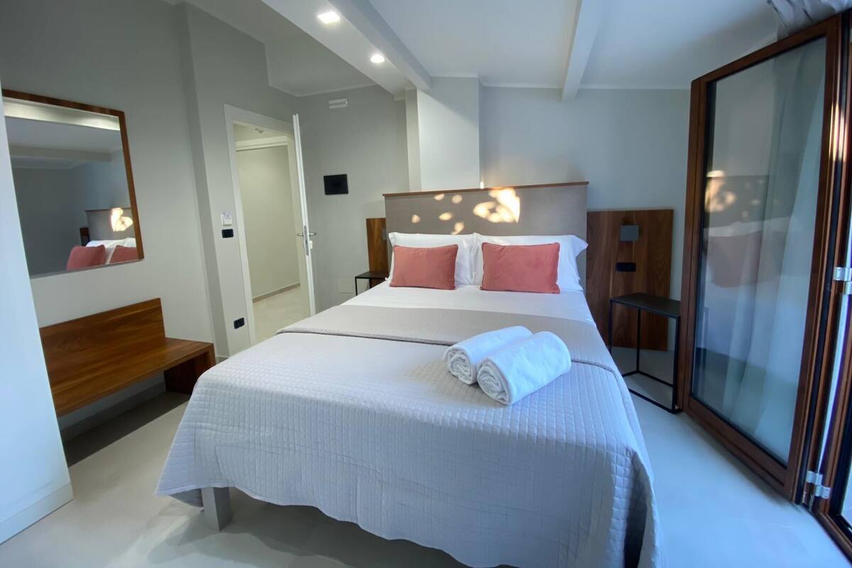 Une chambre d’hôtel moderne avec un grand lit, une table de chevet, un miroir et une porte-fenêtre coulissante.