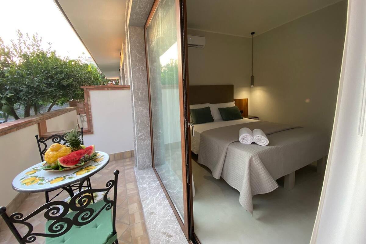 Un balcon avec une table dressée, ornée de fruits, et une chaise, un lit avec une tête de lit et une table de chevet avec une lampe.