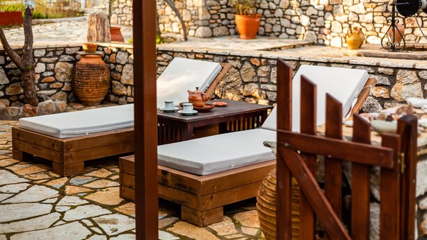 A stone-floored outdoor seating area with wooden lounge chairs and a table, surrounded by stone walls and potted plants. - The Fairytale House Seaside Retreat (Messini)