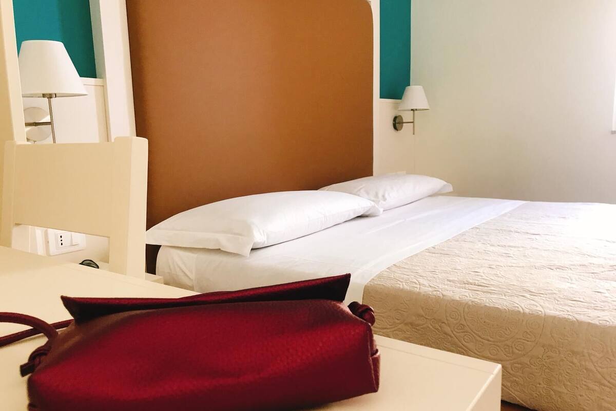 A hotel room with a bed, white headboard, two bedside lamps, and a red bag on a table.