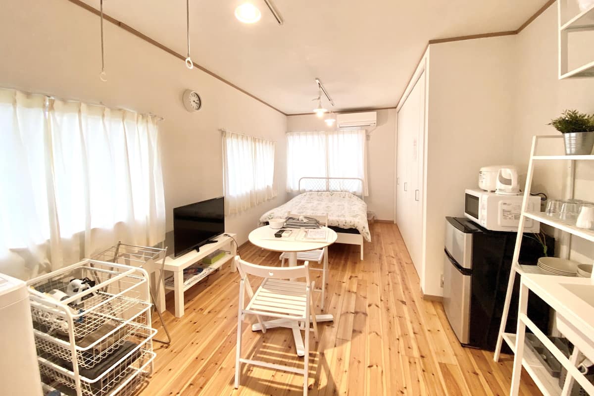 A compact room with a bed, a small table, a chair, a TV, a microwave, and a shelf with items.