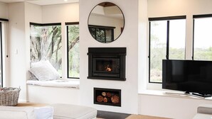 A modern living room with a fireplace, a television, and large windows offering a view of trees.
