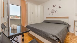 A bedroom with a bed, a desk with two cups, a wardrobe, and a view of buildings through a window. - Apartments Anita Summer (Vodice)
