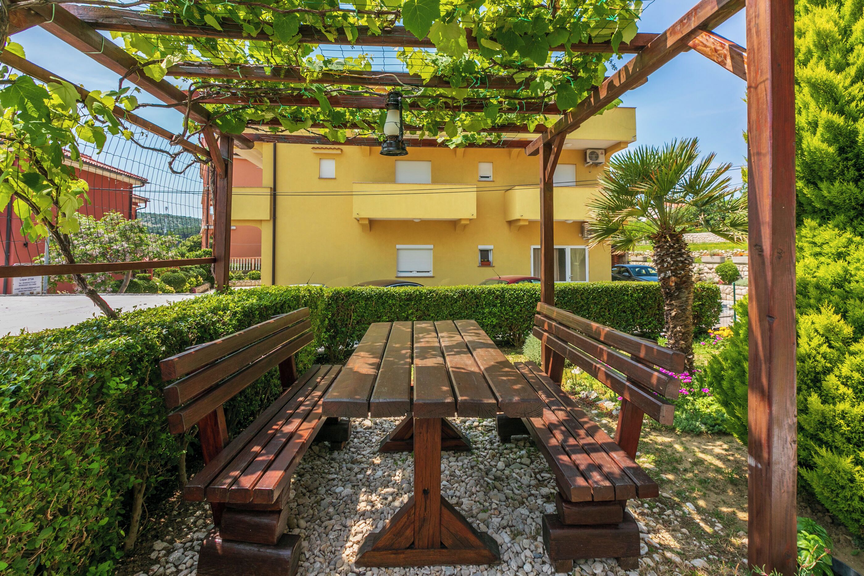 An outdoor wooden bench and table set up under a pergola with a yellow building in the background.