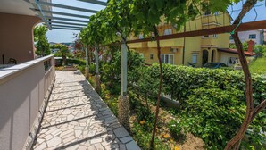 Un sentier pavé de pierres mène à une zone abritée, surmontée d’une pergola, et est entouré d’une végétation luxuriante. Un bâtiment jaune se dresse en arrière-plan.