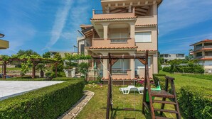 Un immeuble résidentiel à plusieurs étages doté d’un balcon, d’un espace jardin avec une balançoire et surplombant un ciel bleu dégagé.