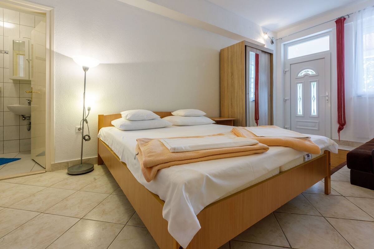 A bedroom with a wooden bed, white linens, a bedside table, a floor lamp, and a view of the outside through a window with curtains.
