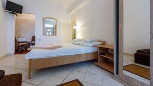 A modern hotel room with a wooden bed, a TV mounted on the wall, a small seating area, and a bathroom visible through an open door. - Apartments Lory (Dramalj)