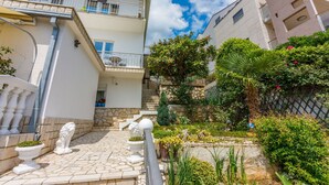 A courtyard with a stone floor, white statues, and a small garden with various plants and flowers. - Apartments Lory (Dramalj)