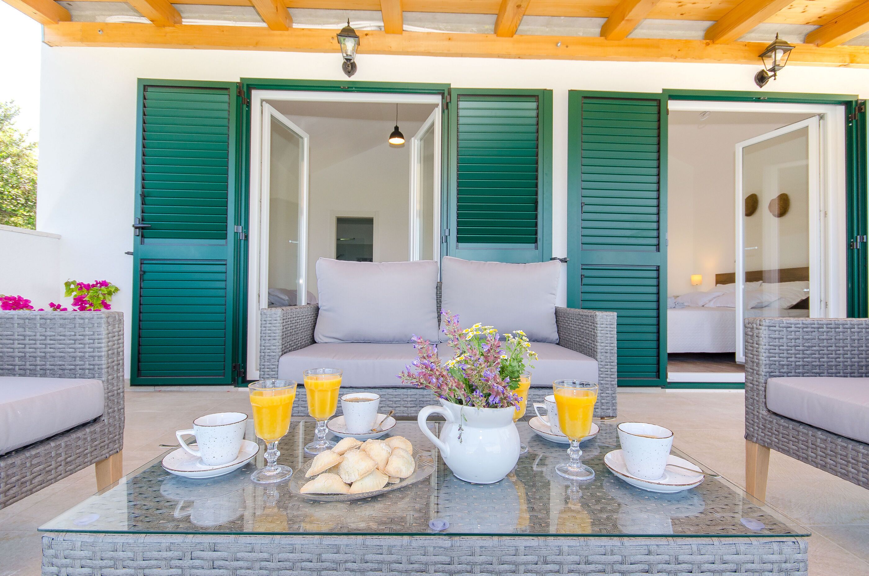 Un área de estar al aire libre con mobiliario de mimbre, una mesa de vidrio y una disposición para el desayuno con cruasanes, jugo y café.
