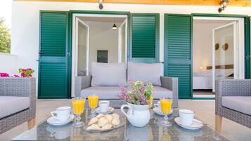 Un área de estar al aire libre con mobiliario de mimbre, una mesa de vidrio y una disposición para el desayuno con cruasanes, jugo y café.