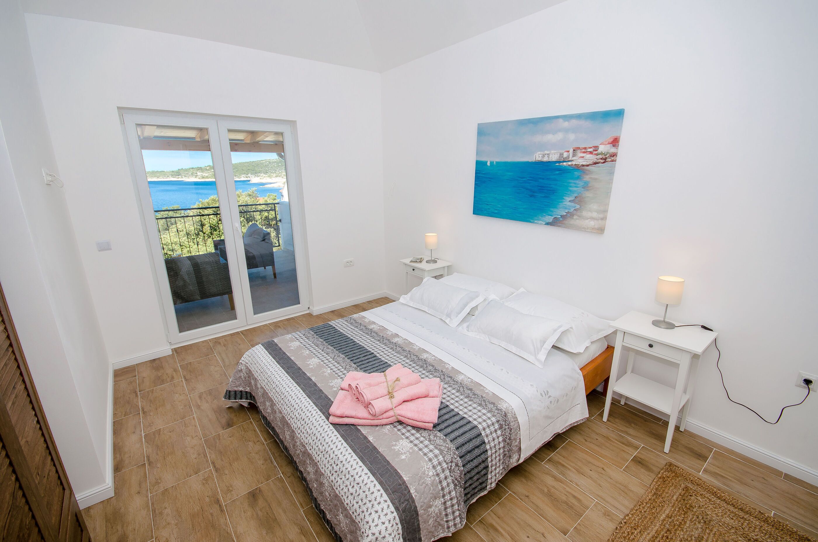 Une chambre à coucher avec un lit, un tableau représentant un paysage côtier, une table de chevet avec une lampe, et une vue sur un espace extérieur accessible par une porte coulissante.