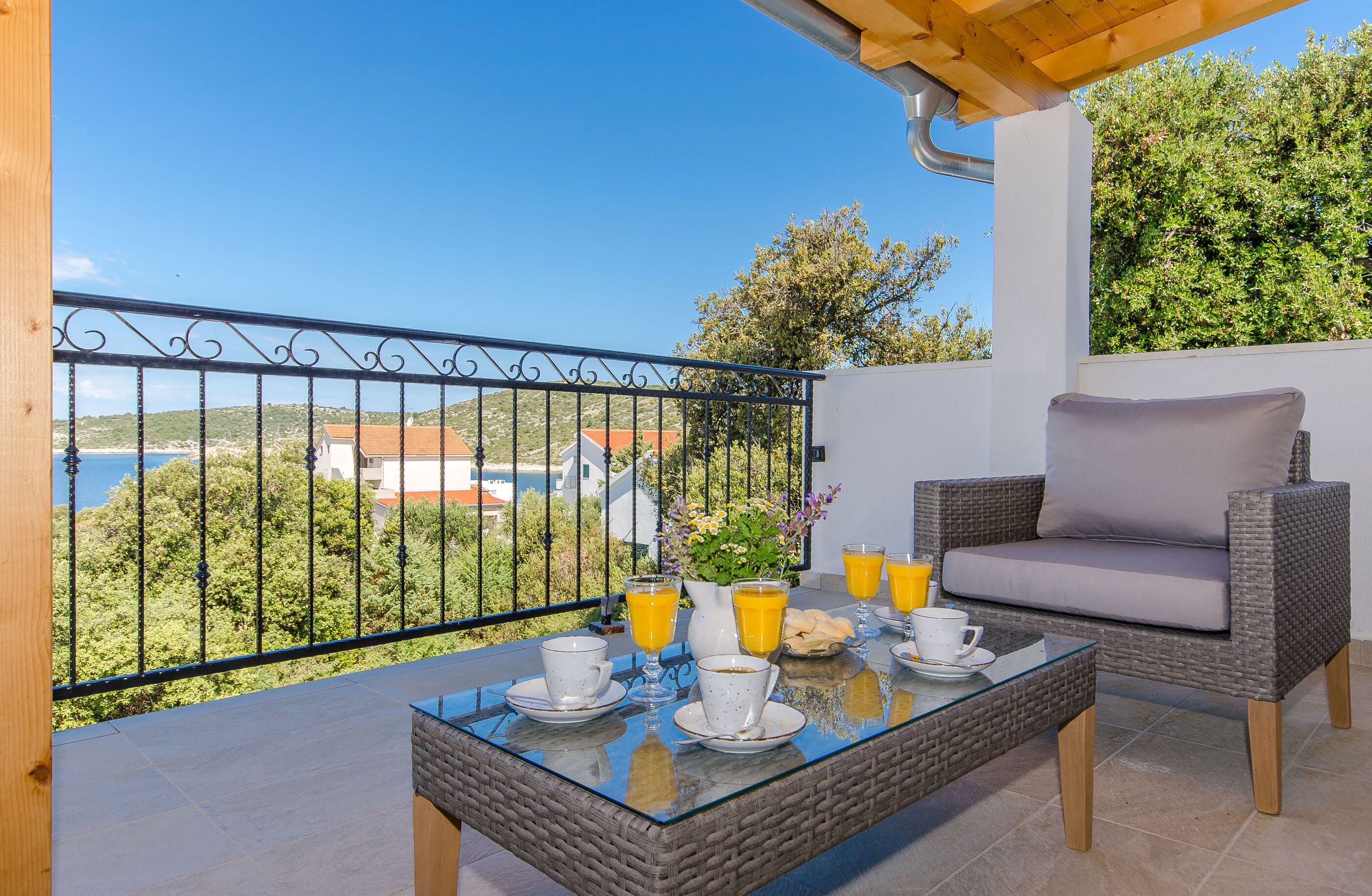 Un balcón con sofá de mimbre y una mesa de vidrio dispuesta con alimentos para el desayuno, con vistas a un paisaje pintoresco.